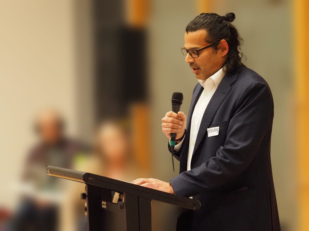 Hitendra Joshi speaking into a microphone at a podium at a community forum