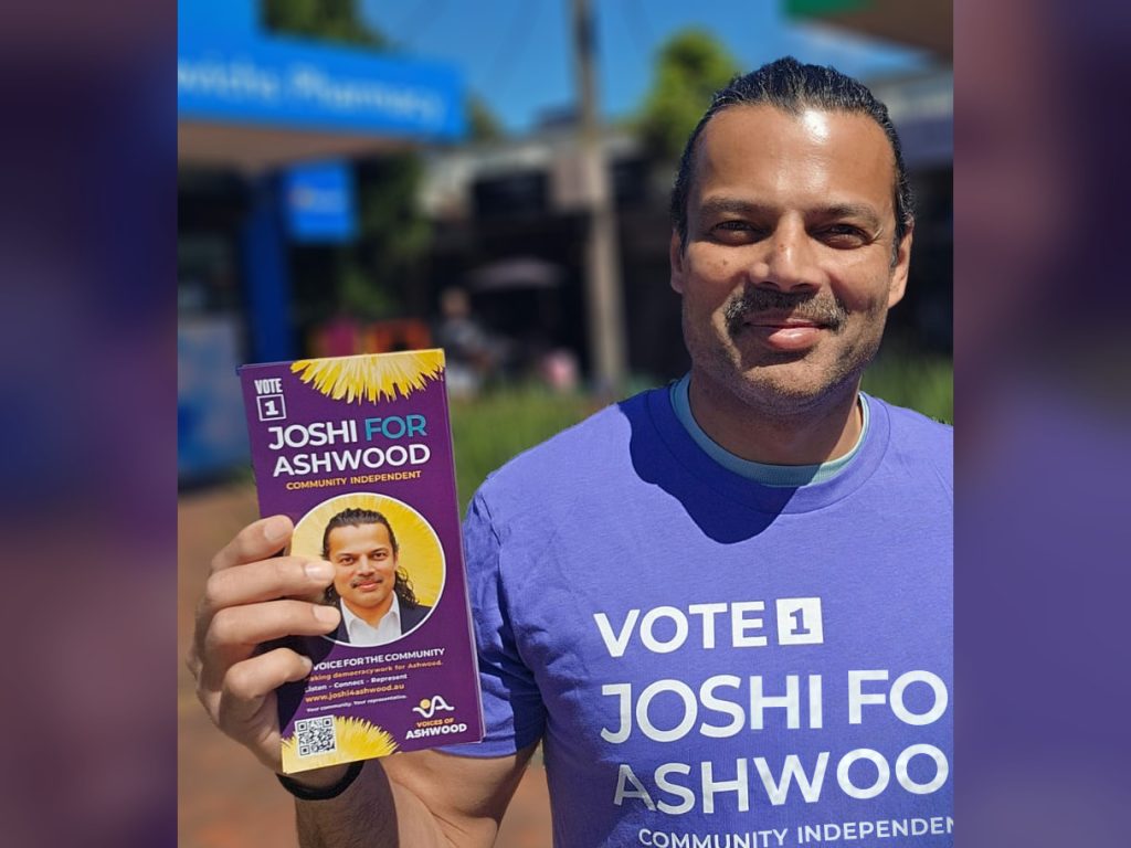 Hitendra Joshi smiling on an Ashwood shopping strip, holding up a Joshi for Ashwood flyer and wearing a purple Vote 1 Joshi for Ashwood t-shirt