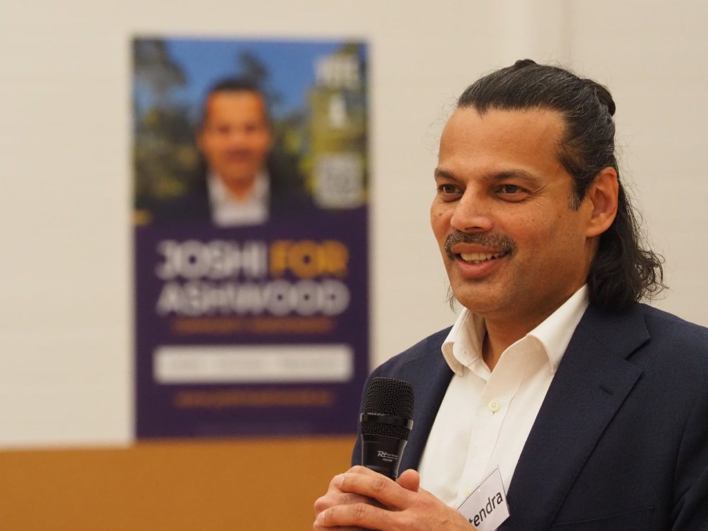 Hitendra Joshi smiling as he speaks into a microphone at a campaign event, with a Joshi for Ashwood poster behind him