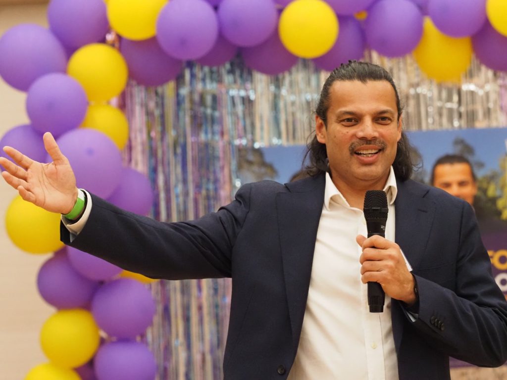 Hitendra Joshi, microphone in hand and arm outstretched, addressing supporters in front of purple and yellow balloons at a campaign event